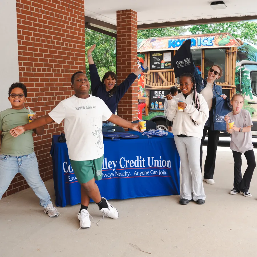 Group of kids and adults enjoying drinks near a Kona Ice truck and a credit union promotional table outdoors.