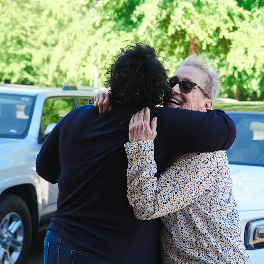 Two people warmly embracing outdoors near parked cars with green trees in the background on a sunny day.