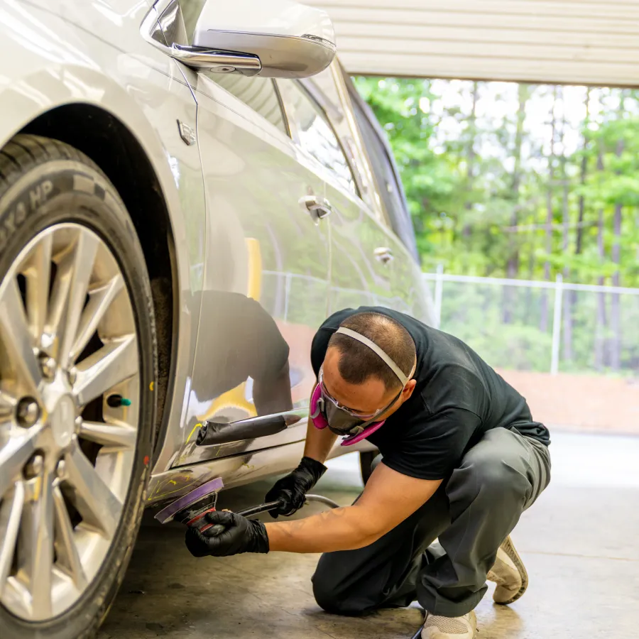 Man wearing protective mask polishing silver car door with electric buffer in garage workshop.