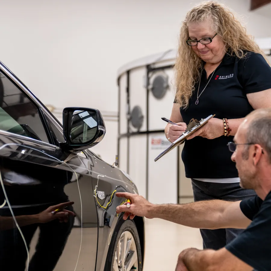 Man inspecting a car door with a tool while a woman takes notes on a clipboard in a garage.