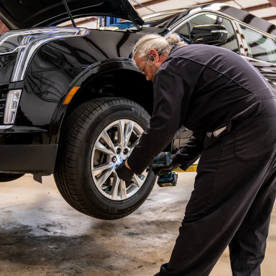 Mechanic in black uniform fixing a tire on a lifted black SUV inside a brightly lit garage.
