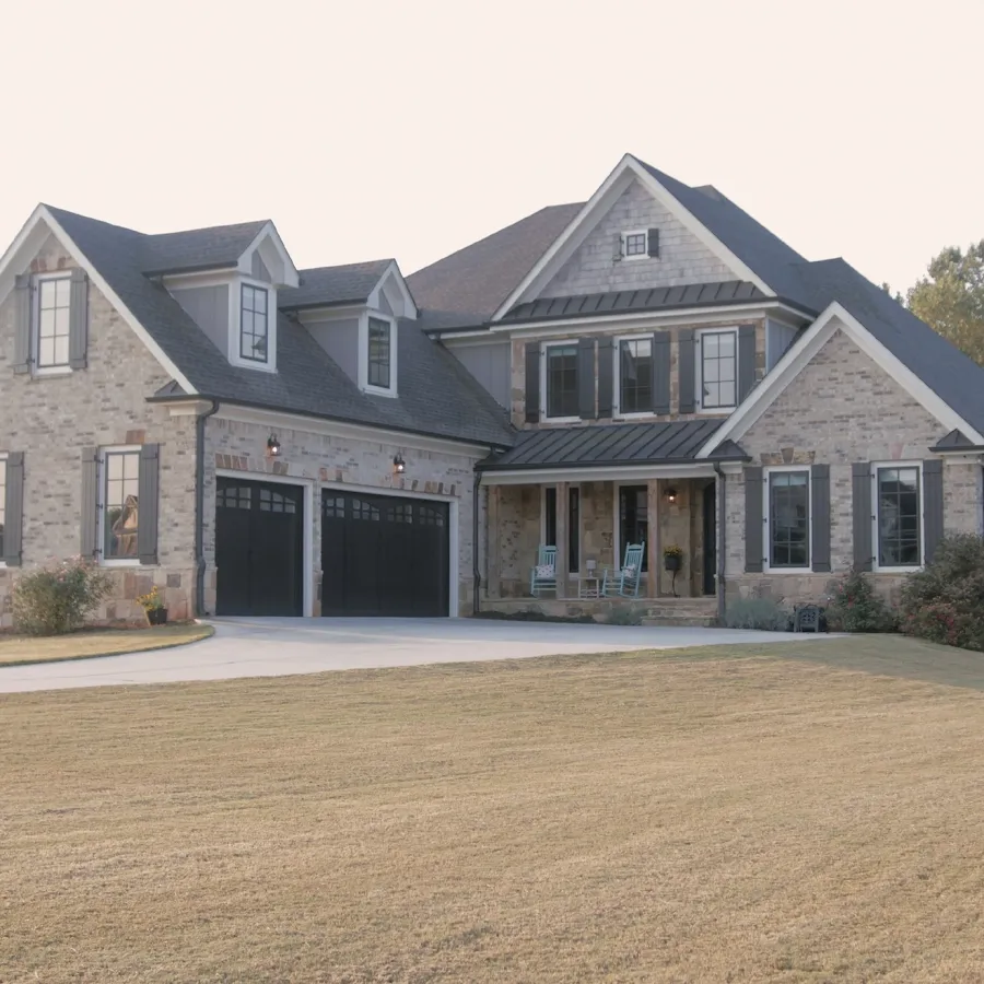 Large modern stone house with dark roof, black garage doors, and a wide driveway surrounded by a spacious lawn.
