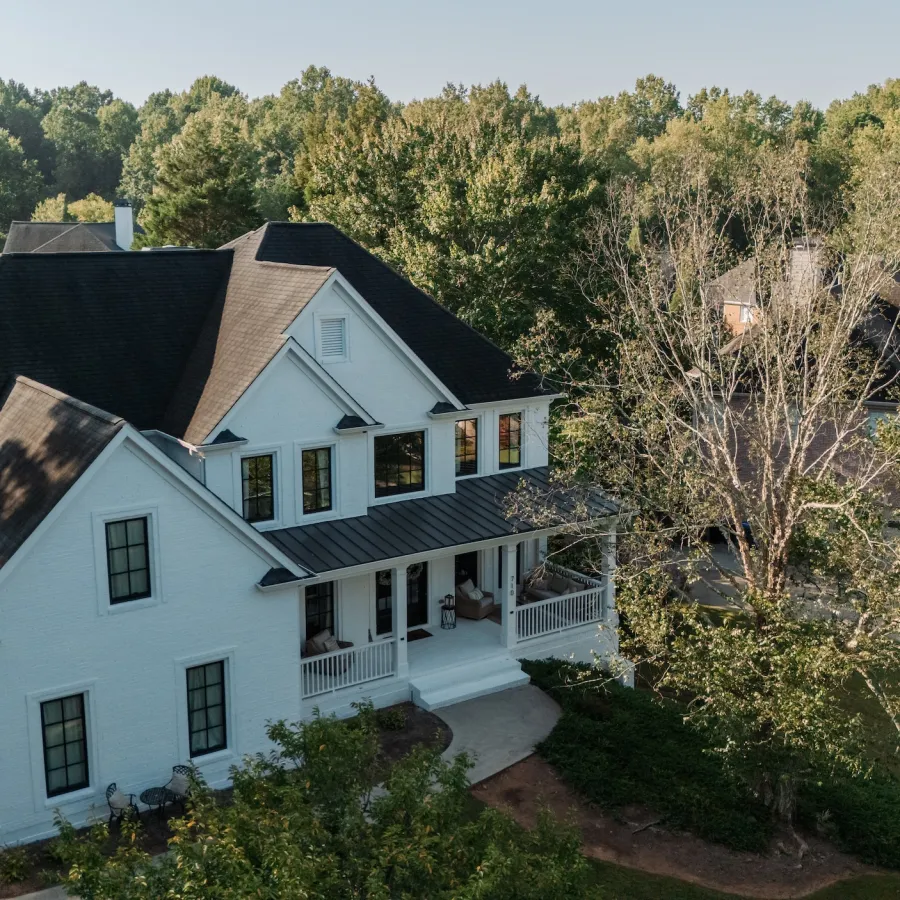 Large white two-story house with black roof surrounded by trees and greenery on sunny day