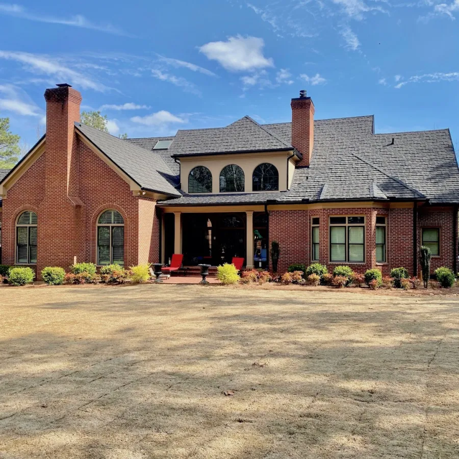 Large brick house with chimneys, arched windows, porch with red chairs, and a spacious dry lawn under blue sky.