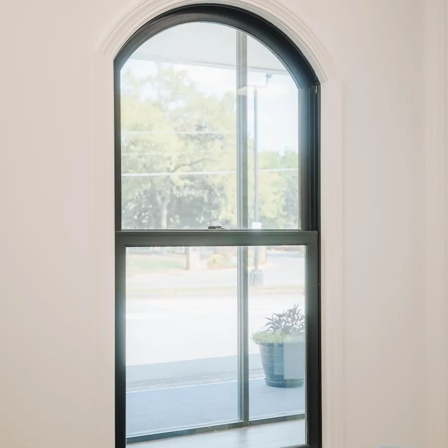 Arched black window with clear glass showcasing an outdoor plant pot and street view inside a white room.
