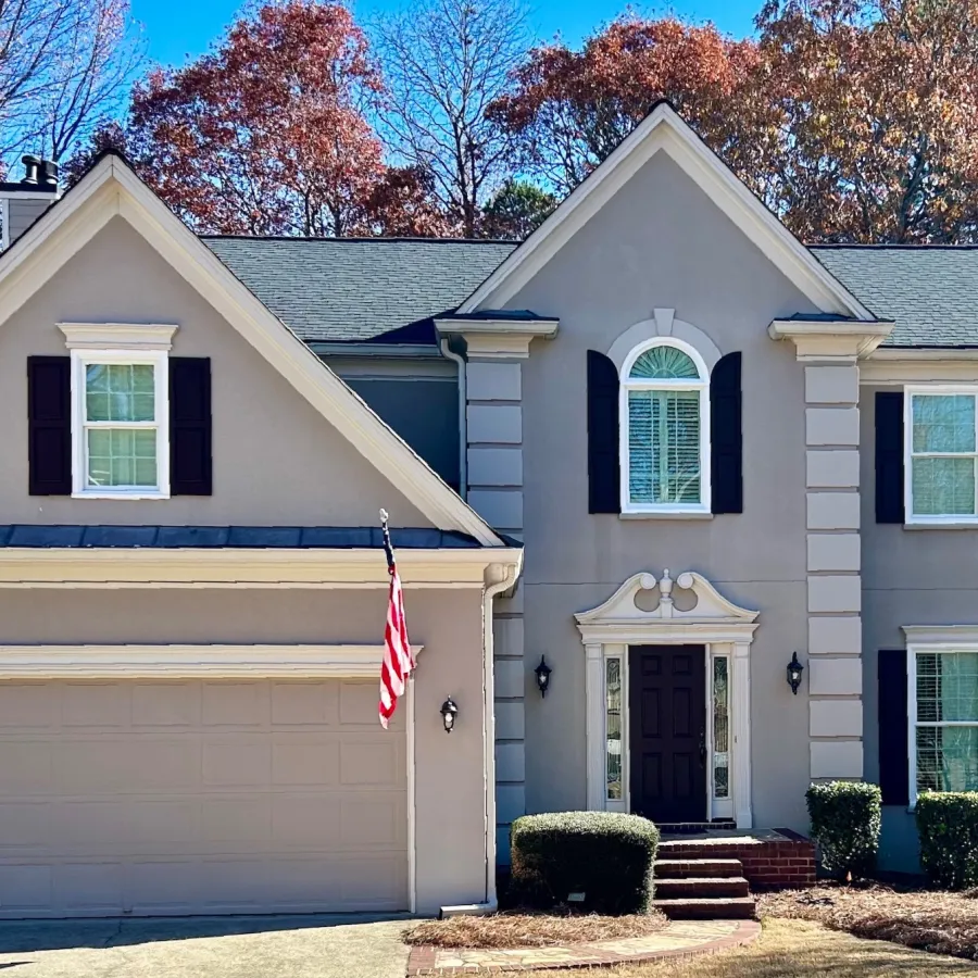 Two-story suburban house with beige exterior, black shutters, attached garage, and American flag in front yard