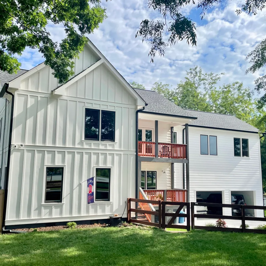 Modern two-story white home with large windows and wooden deck, surrounded by greenery.
