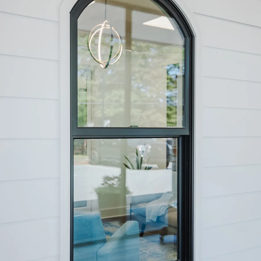 Arched black-framed window on white siding reflects interior with blue chairs and modern light fixture.