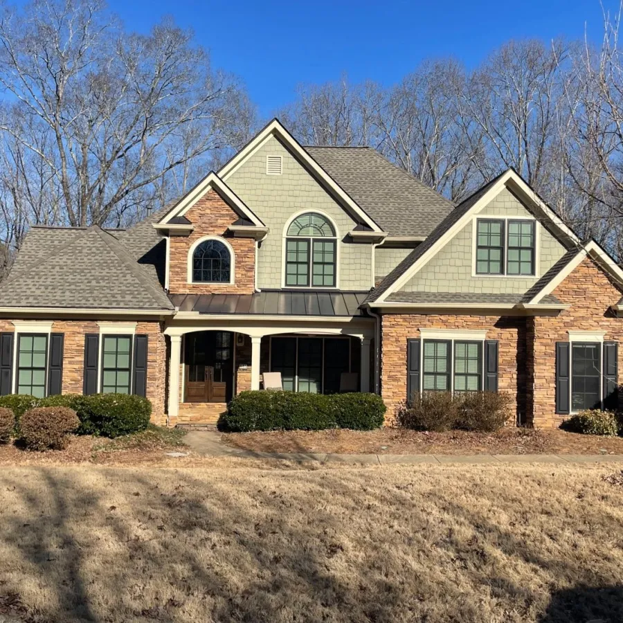 Large two-story house with grey roof, brick and siding exterior, symmetrical windows, and front porch under clear blue sky.
