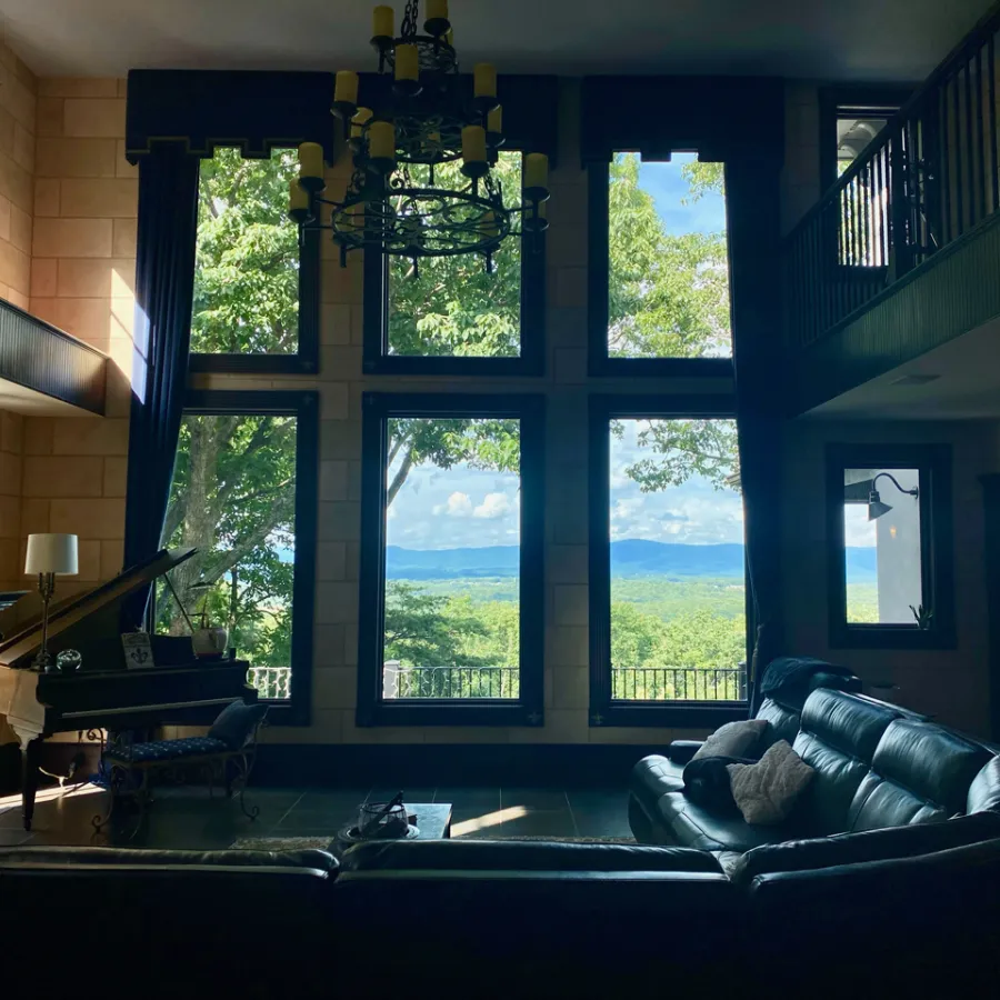 Spacious living room with large windows overlooking lush green trees and distant mountains under a blue sky.