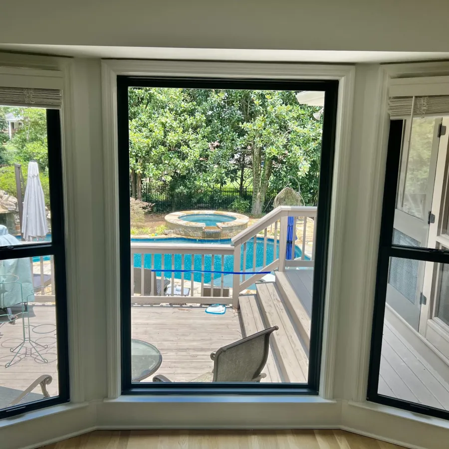 View through three black-framed windows of backyard deck, pool, hot tub, and lush green trees on a sunny day