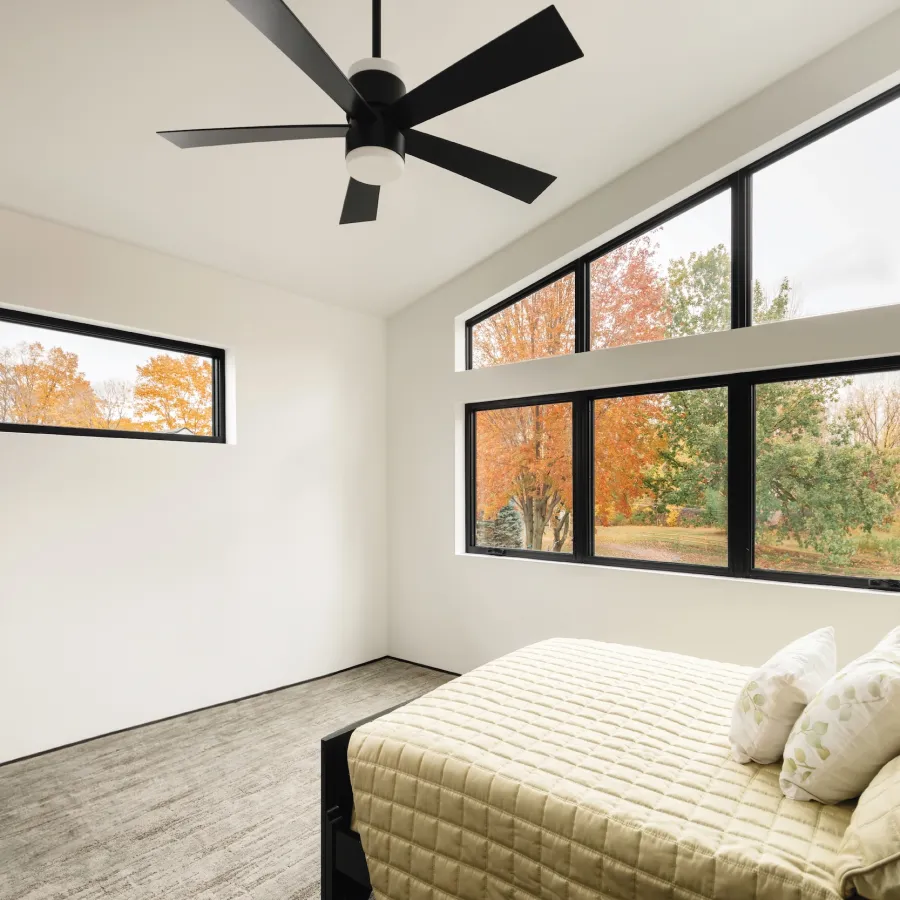 A bedroom with a black ceiling fan and custom shape windows overlooking the yard and trees outside