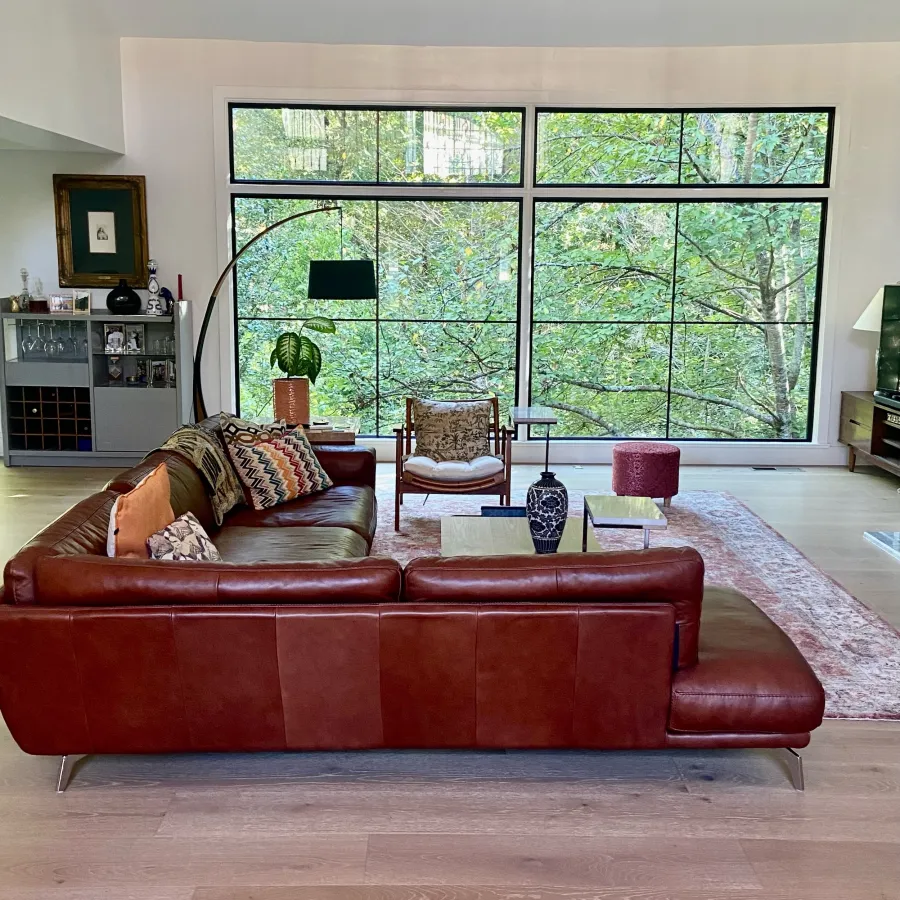 Modern living room with large fiberglass windows, brown leather sofa, stylish decor, and greenery outside.