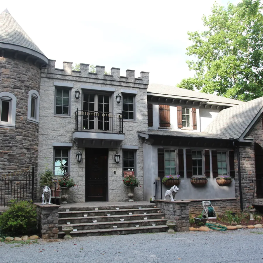 Stone castle-style house with turret, steps, flower pots, and lush green trees in the background