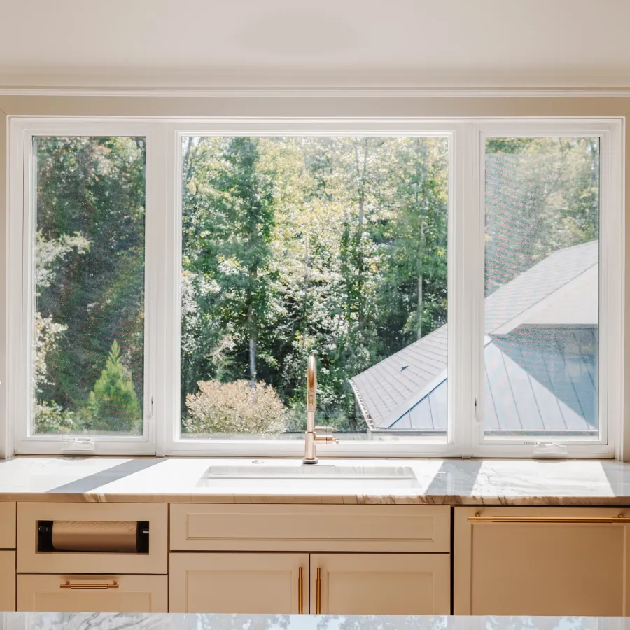 Bright kitchen sink with marble countertop and large window showing green trees outside.