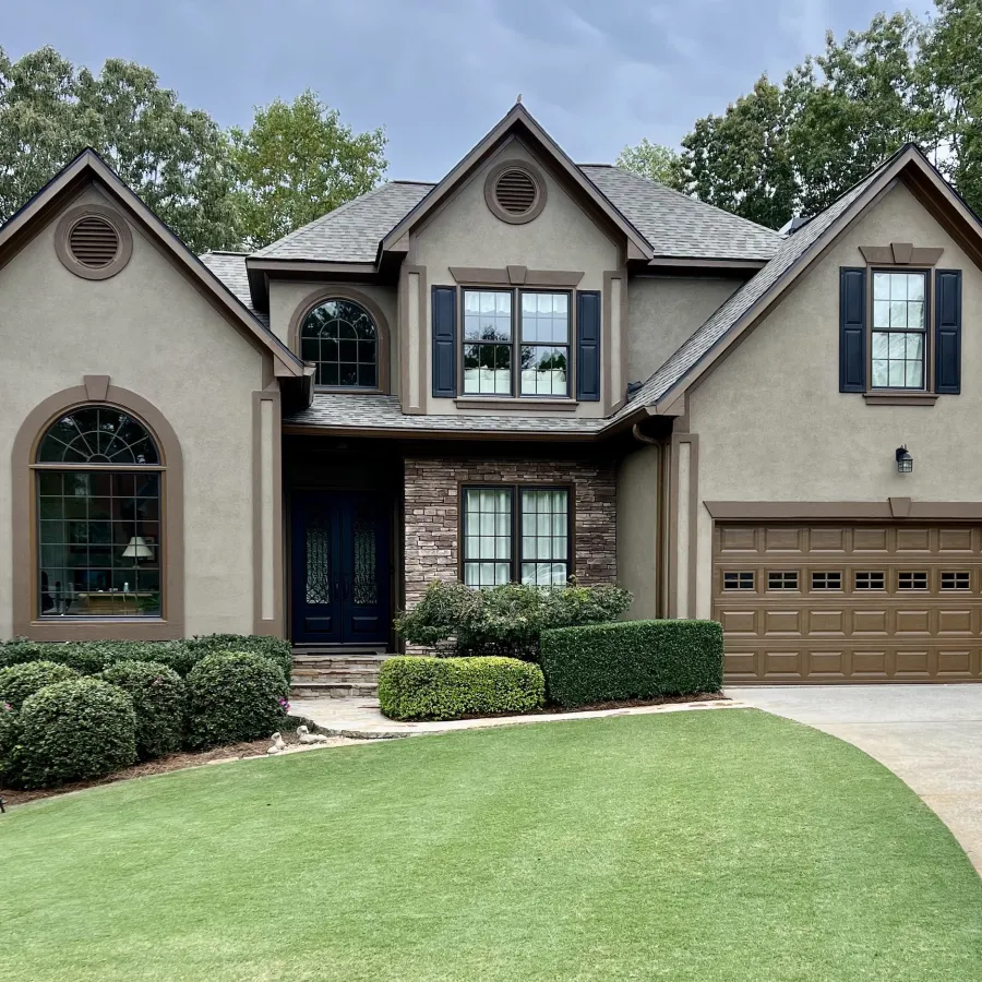 Two-story beige house with brown accents, large windows, manicured lawn, shrubs, and attached garage under trees.