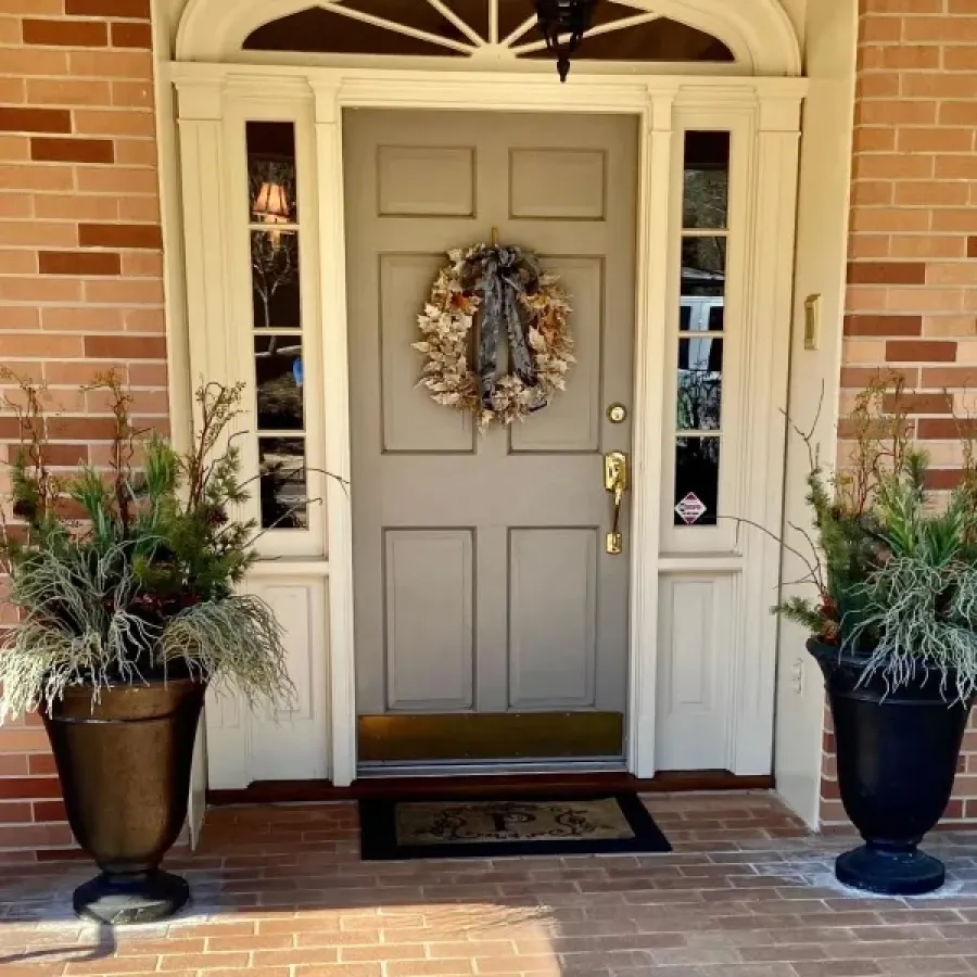 Front gray door with decorative wreath, flanked by black planters with greenery on brick porch under lantern light.