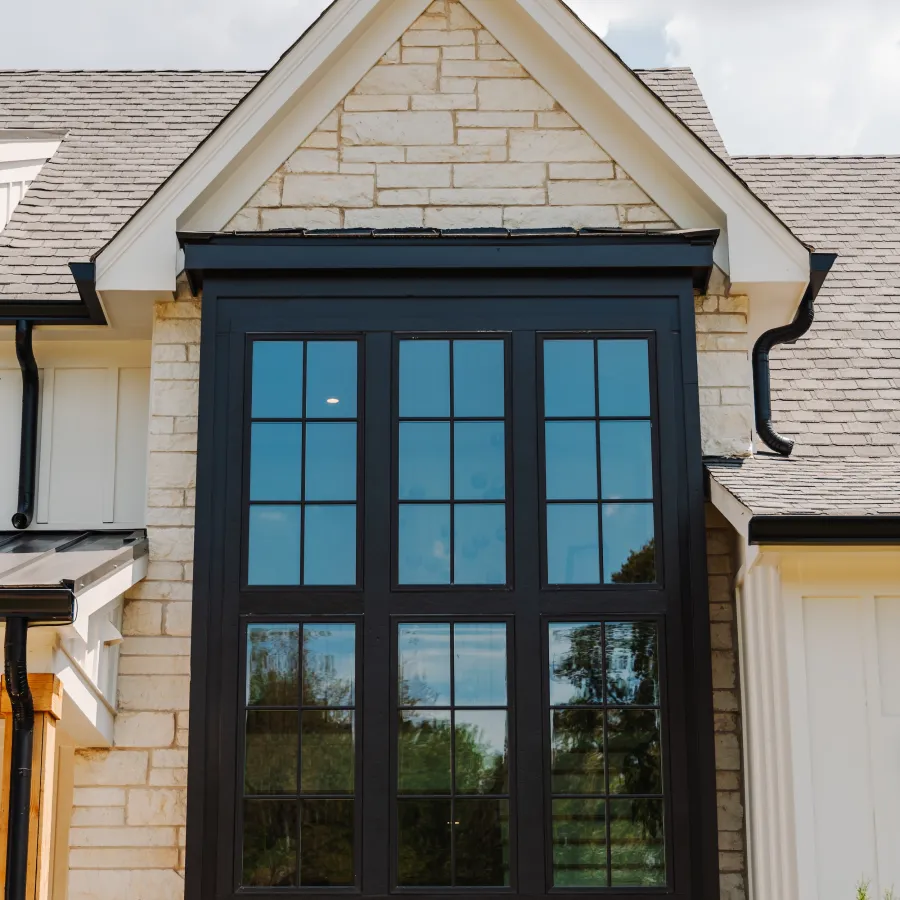 Large black framed window with grid pattern on a beige stone house under a cloudy sky