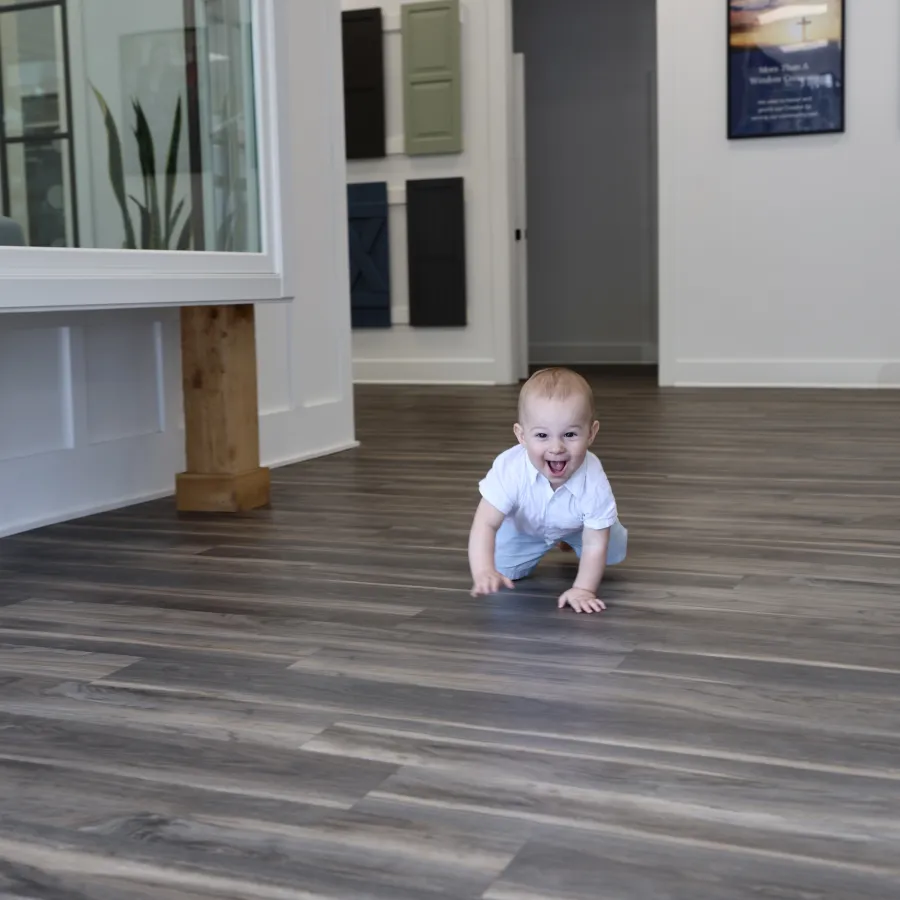 Happy baby crawling on modern wood-look flooring in a bright, spacious room with white walls and decor samples.