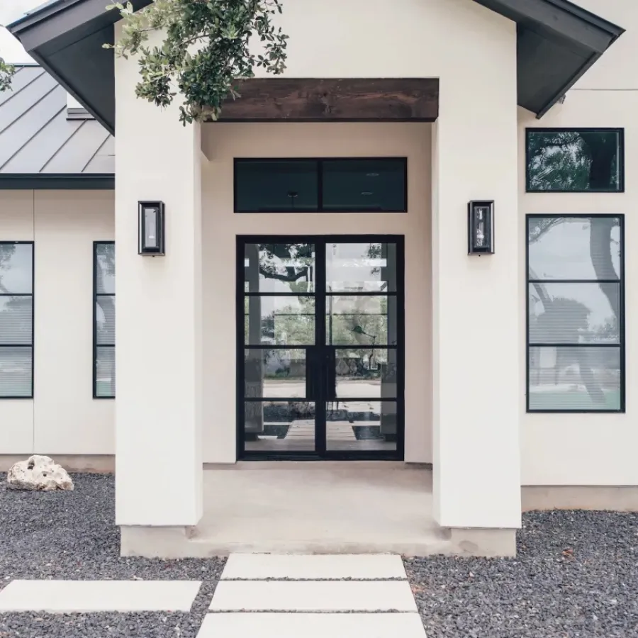 Modern white house entrance with large black-framed glass door and windows, surrounded by gravel and trees.