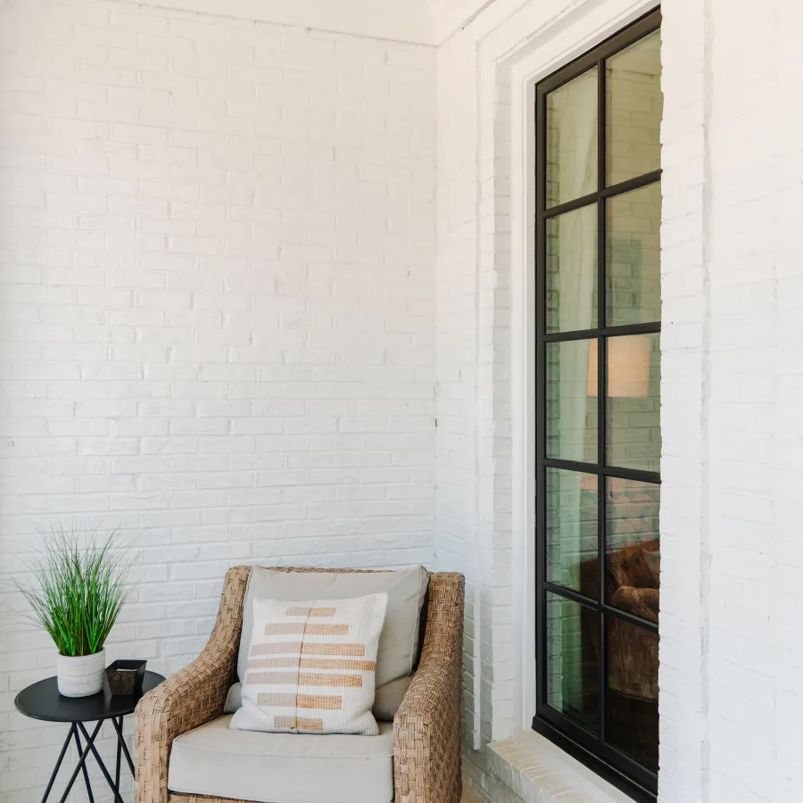 Cozy wicker armchair with cushion beside small round table and potted plant on white brick porch.