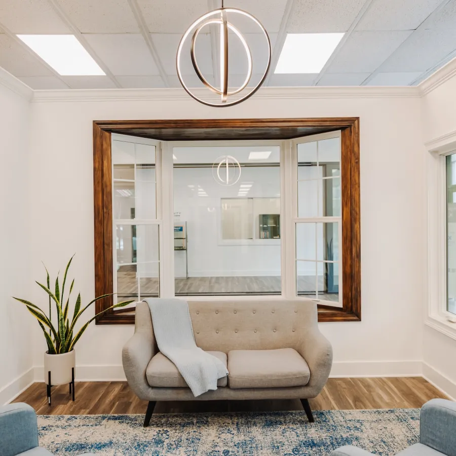 Modern cozy waiting room with a gray sofa, blue rug, wooden floor, indoor plant, and large window reflections.