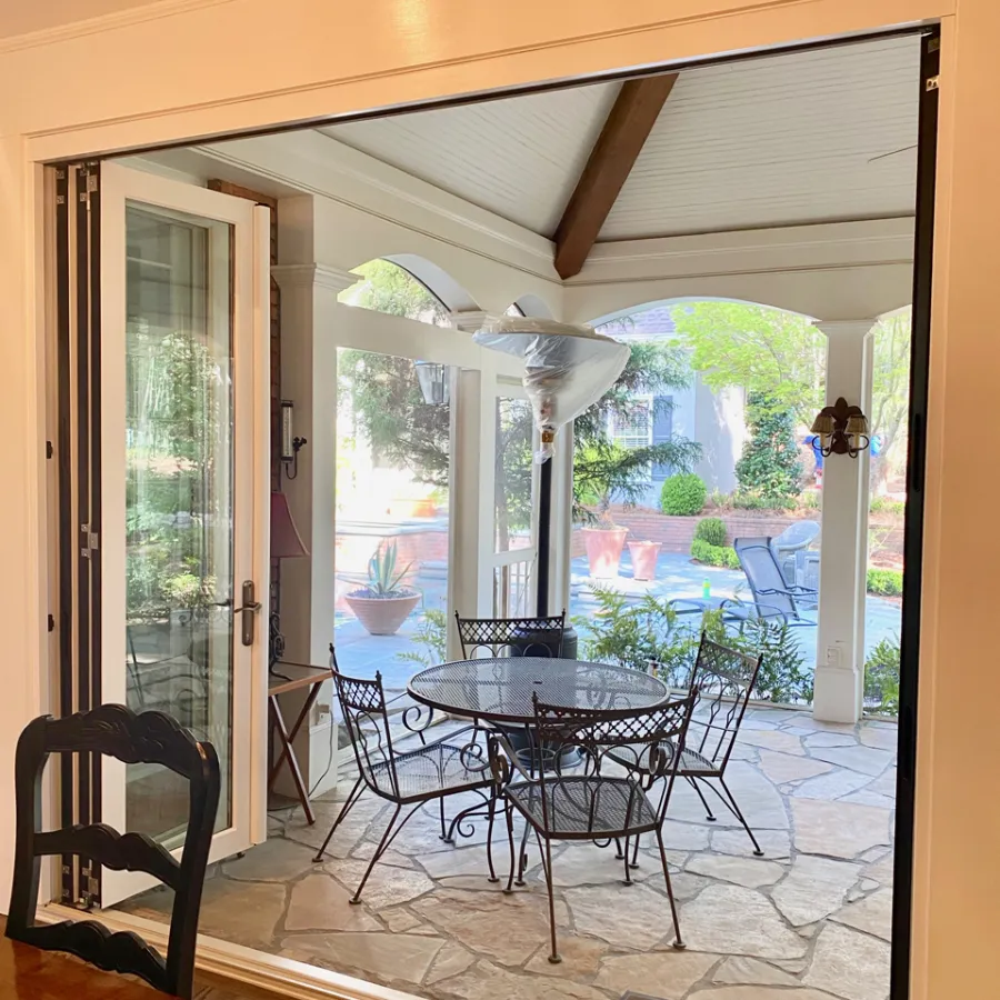 Open folding glass door reveals outdoor patio with metal table and chairs under covered porch with greenery.