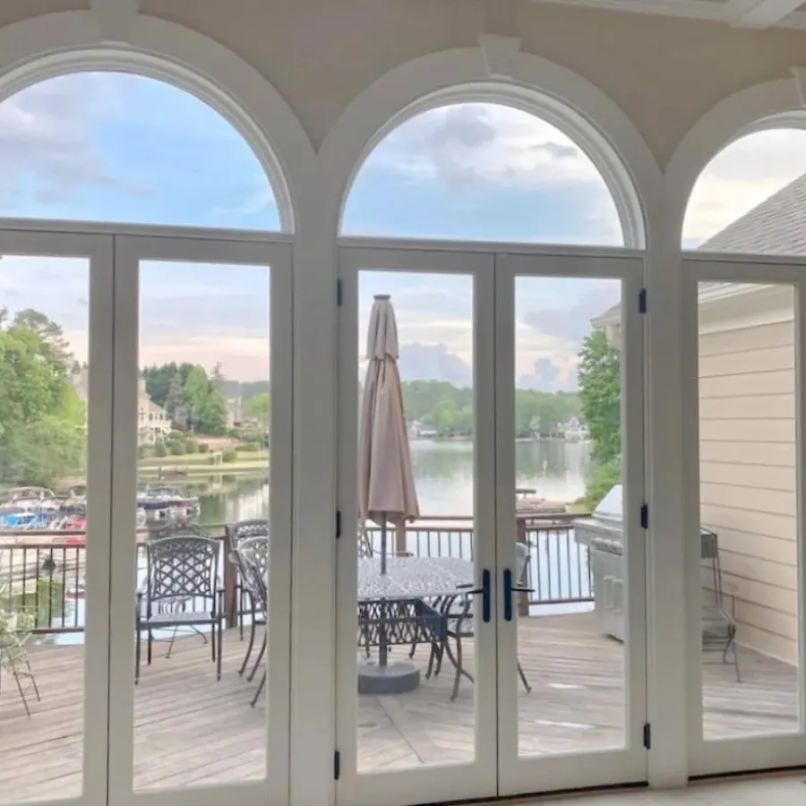 View through arched glass doors onto a lakeside deck with outdoor furniture and umbrella on a sunny day