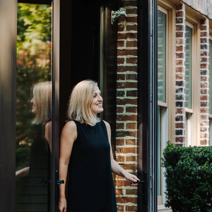 Woman in a black dress smiling and stepping outside through a glass door of a brick house with greenery.