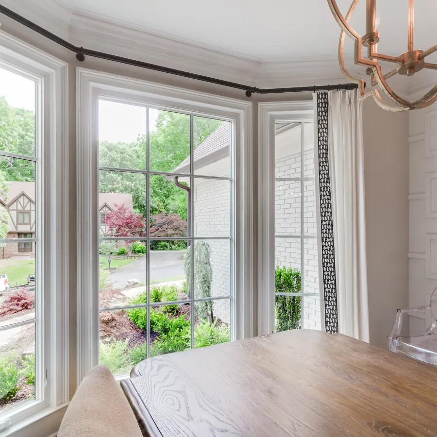Bright dining area with large windows framed by elegant curtains, showcasing a lush garden view.