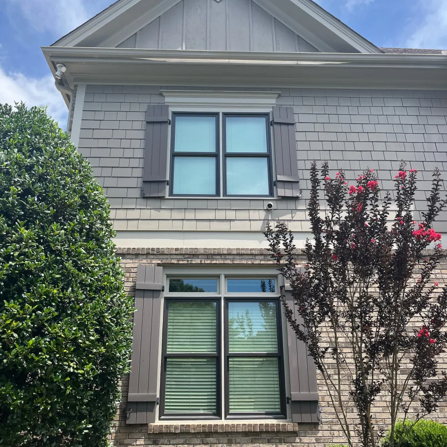 Modern house exterior with brick and siding facade, two windows with shutters, and green landscaping under a blue sky.