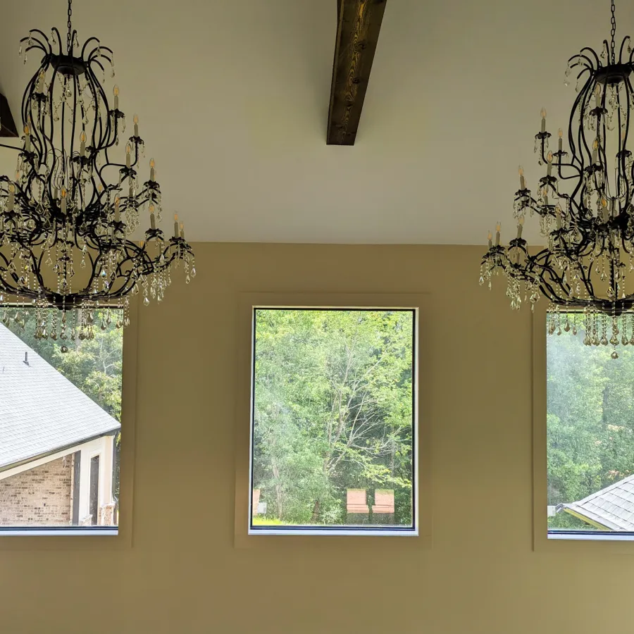 Two ornate black chandeliers with crystals hang in a room with three large square windows showing trees outside.