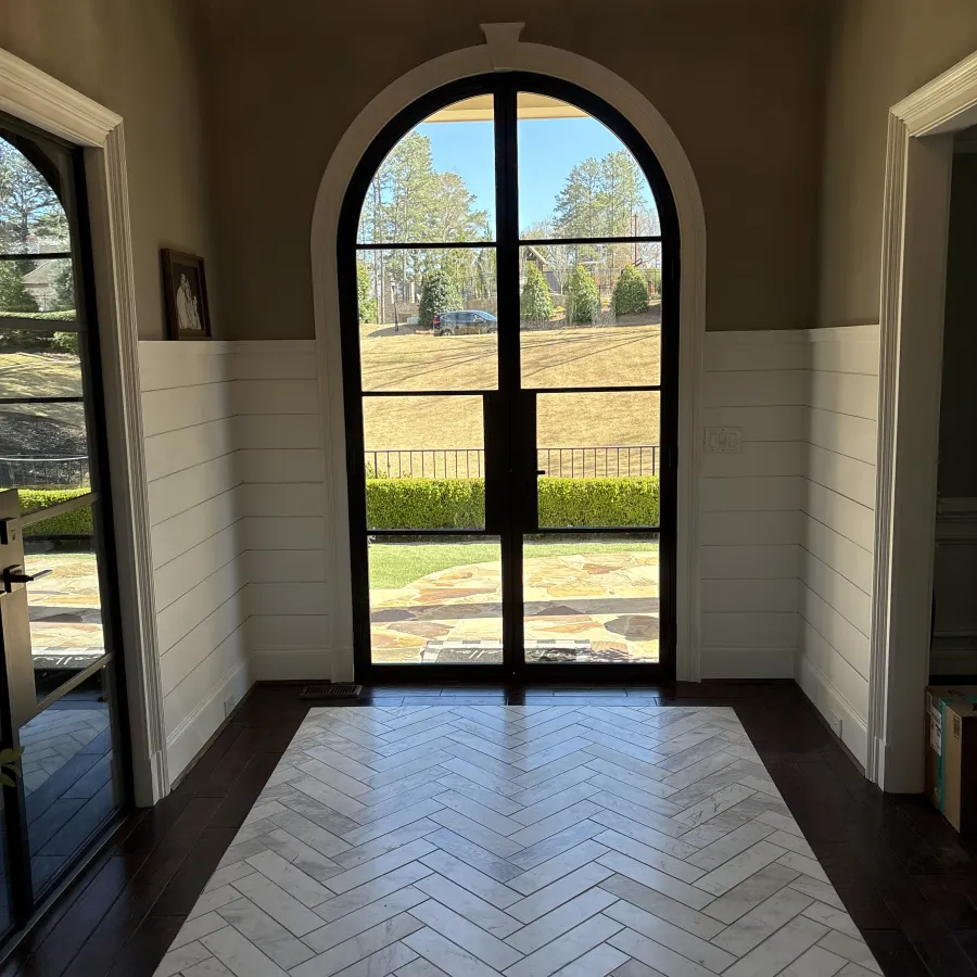 Indoor view of an arched glass door leading to a sunlit backyard with trees and grass.
