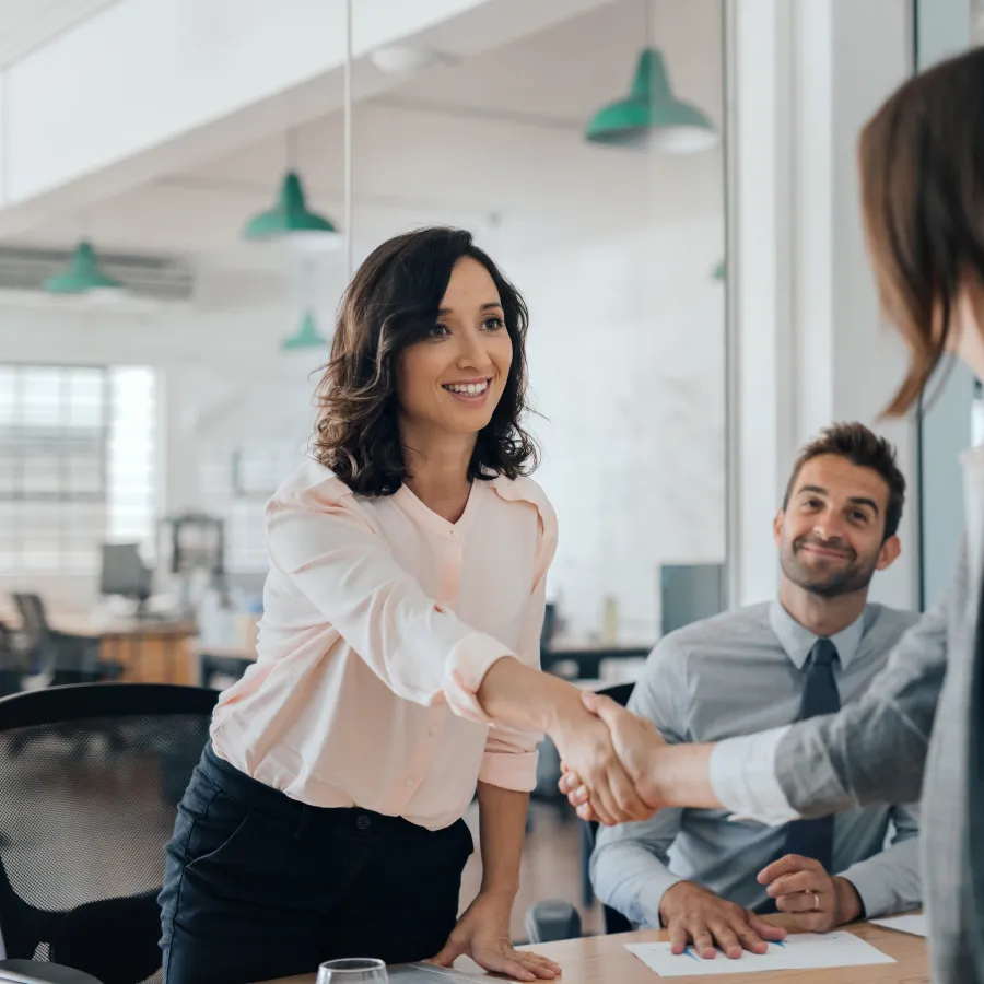 Two businesswomen shaking hands during a meeting with a man smiling in a modern office setting