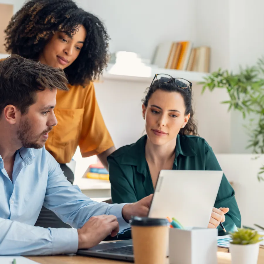Three colleagues collaborate on a laptop in a modern office with plants and shelves in the background