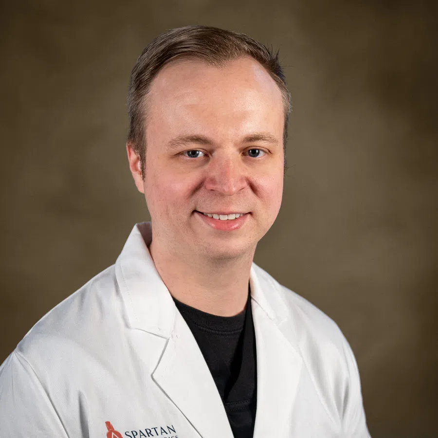 Portrait of a man wearing a white lab coat over a black shirt, smiling against a plain brown background.