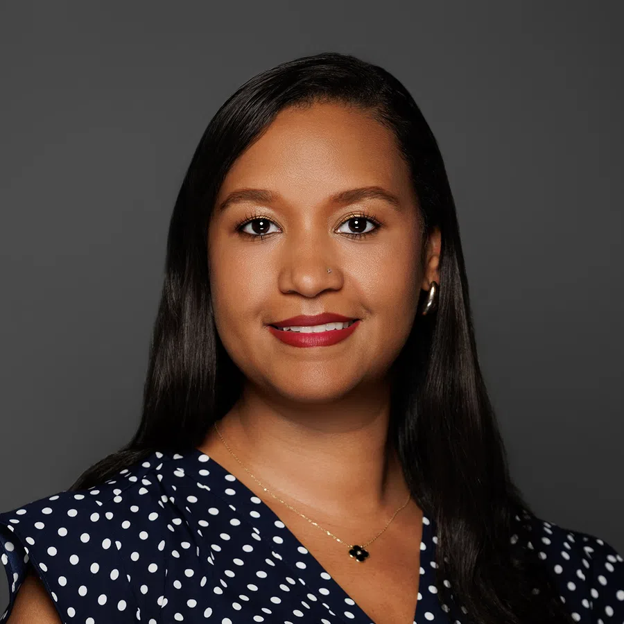 Confident woman with long dark hair wearing a navy blue polka dot blouse and red lipstick smiling against gray background