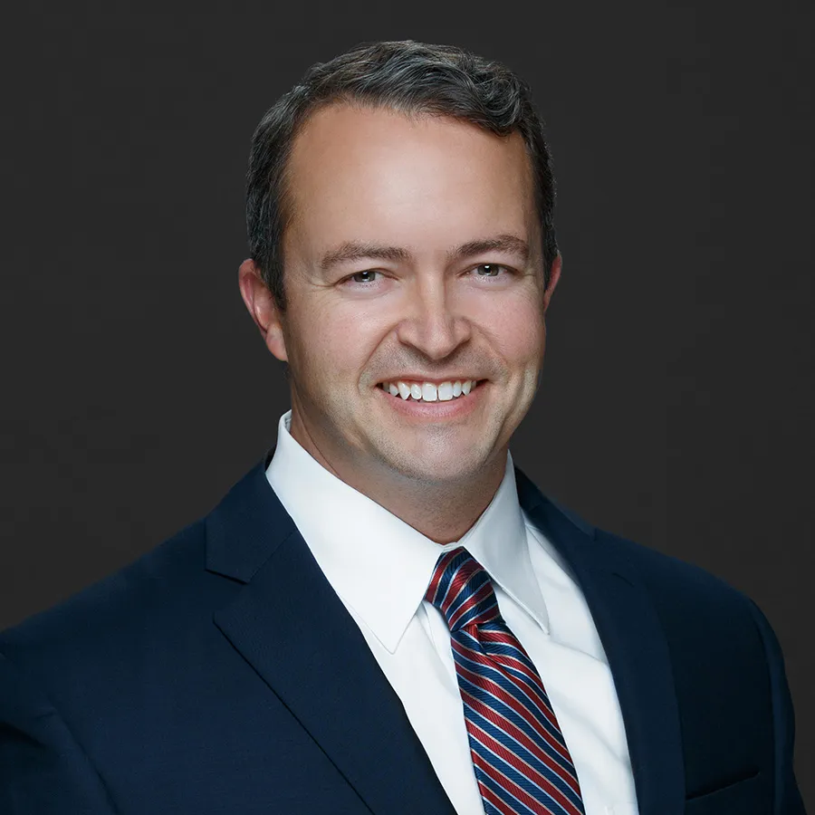 Smiling man in a navy suit, white dress shirt, and red striped tie against a dark background.