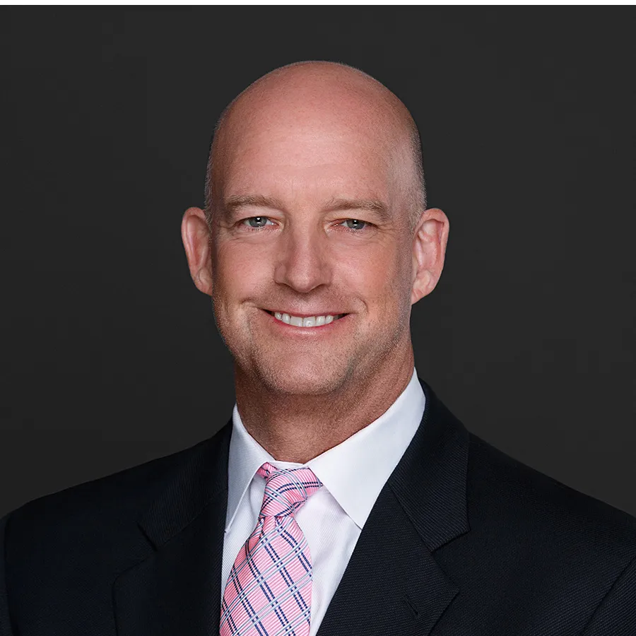 Smiling bald man in a black suit and pink patterned tie against a dark background.