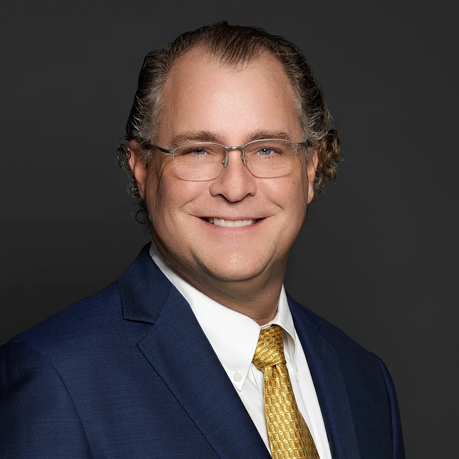 Smiling middle-aged man with glasses in a blue suit and gold tie against a dark gray background
