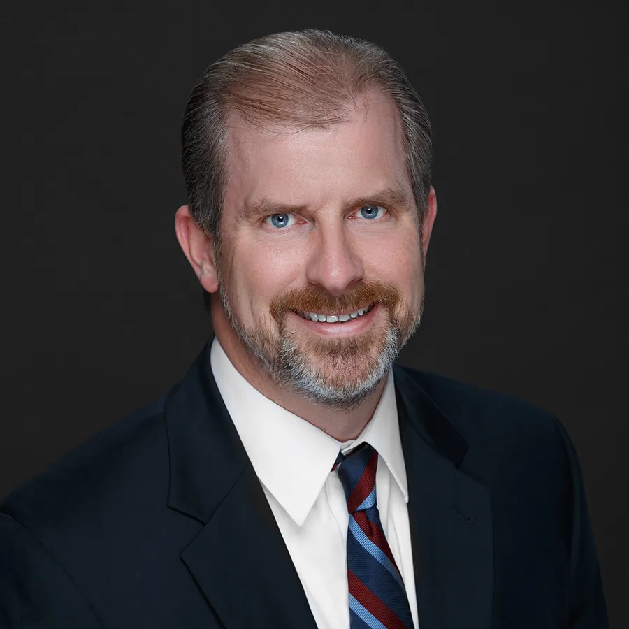 Professional headshot of smiling middle-aged man with beard wearing dark suit and striped tie against black background
