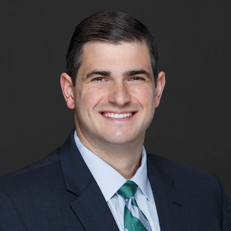 Smiling man in dark suit, light blue shirt, and green striped tie against black background.