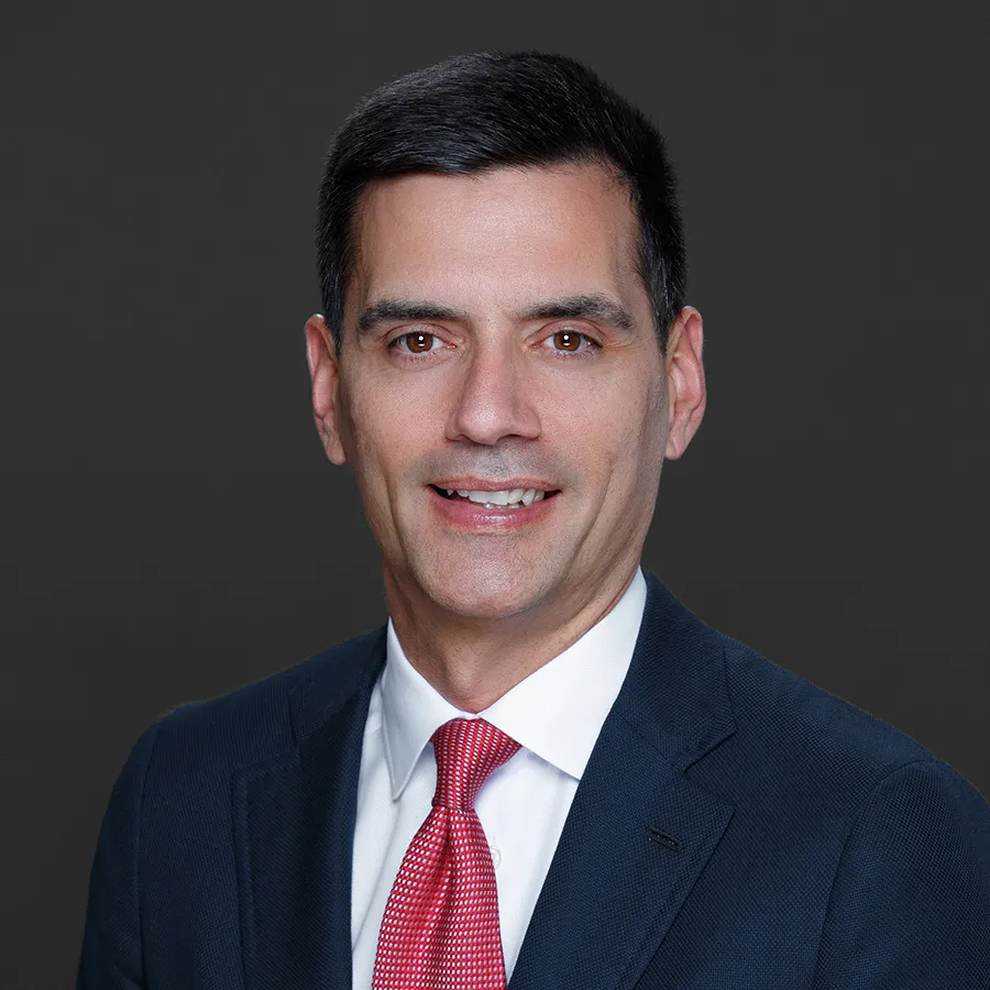 Professional portrait of a man in a dark suit, white shirt, and red tie against a gray background