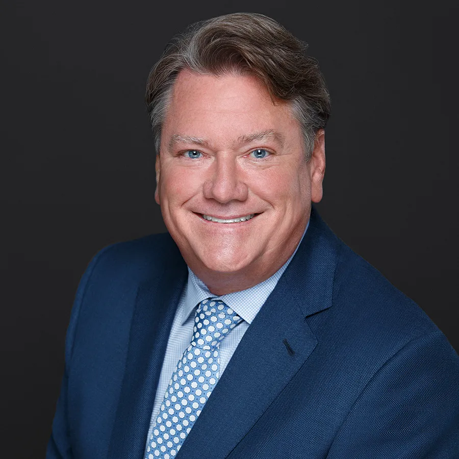 Smiling middle-aged man with blue eyes wearing a blue suit and polka dot tie against a dark background