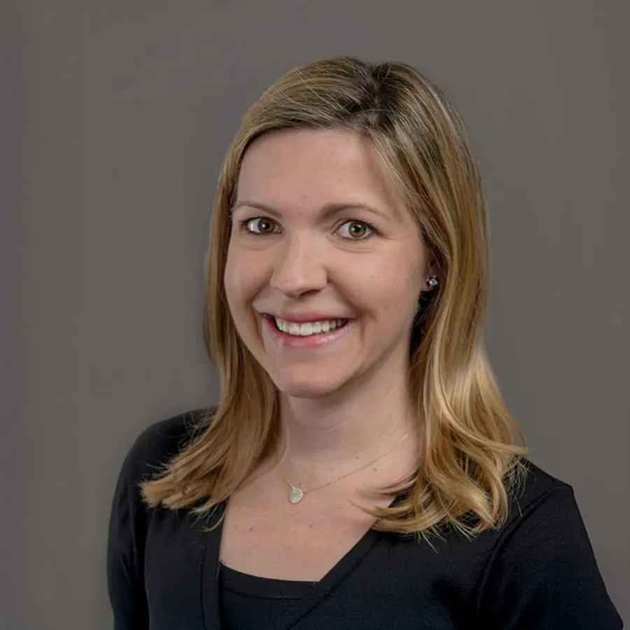 Smiling woman with shoulder-length blonde hair wearing a black top and heart-shaped pendant necklace on grey background