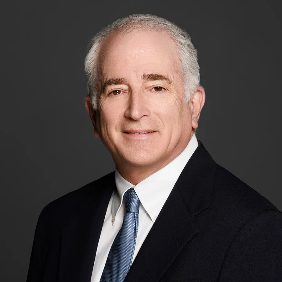 Professional headshot of an older man with gray hair in a dark suit and blue tie against a gray background.