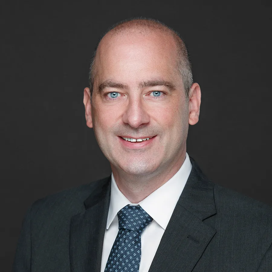 Professional headshot of a smiling bald man in a dark suit and patterned blue tie with a black background