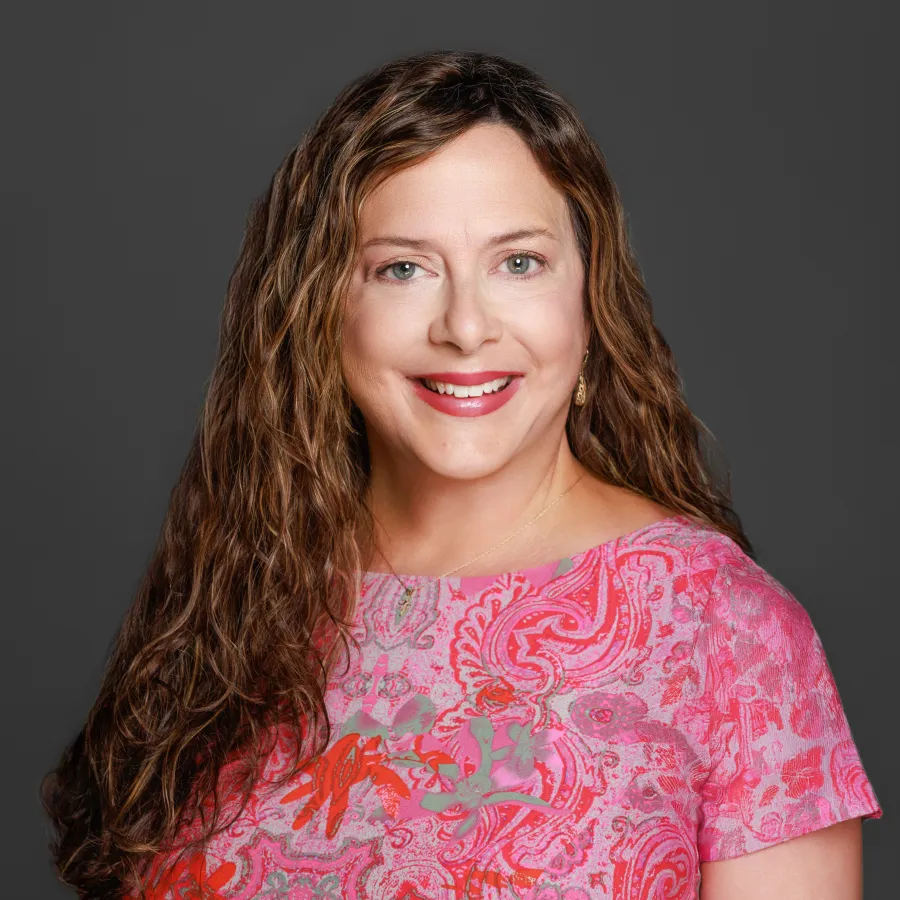 Smiling woman with long, curly hair wearing a vibrant pink paisley top against a gray background.