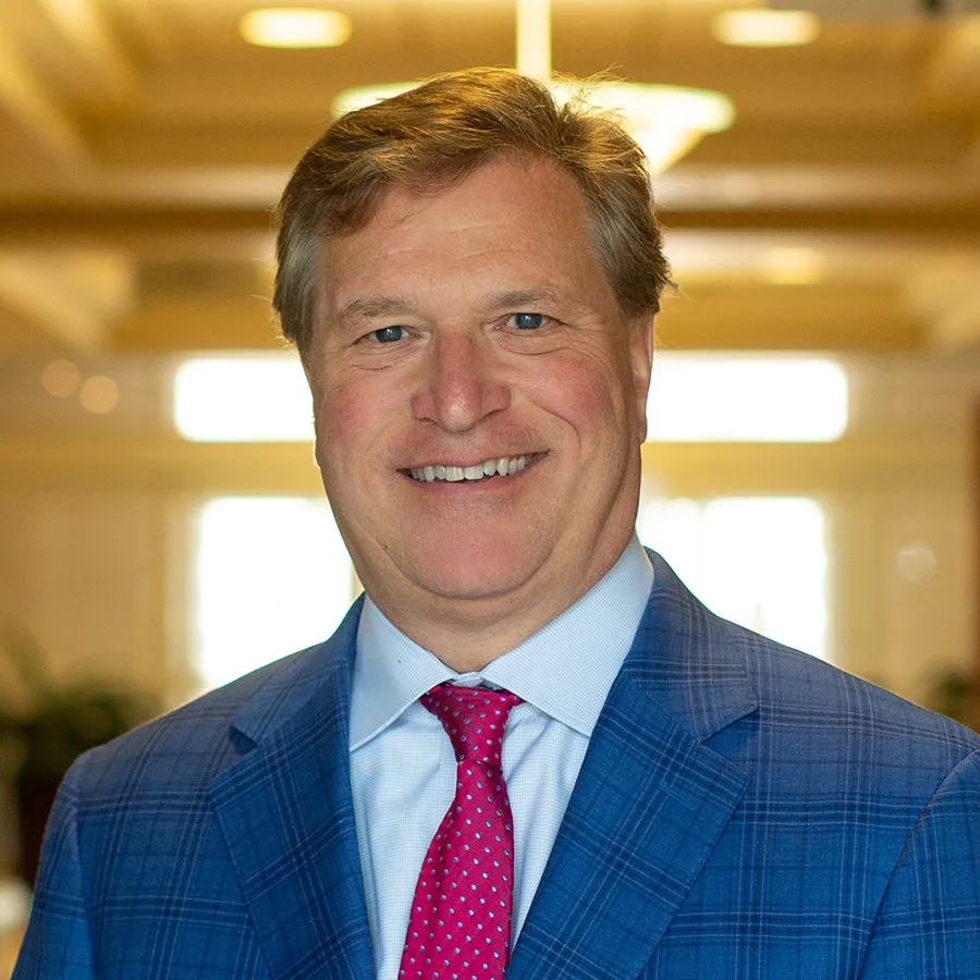 Smiling middle-aged man in a blue checked suit and red tie in a warmly lit indoor setting.