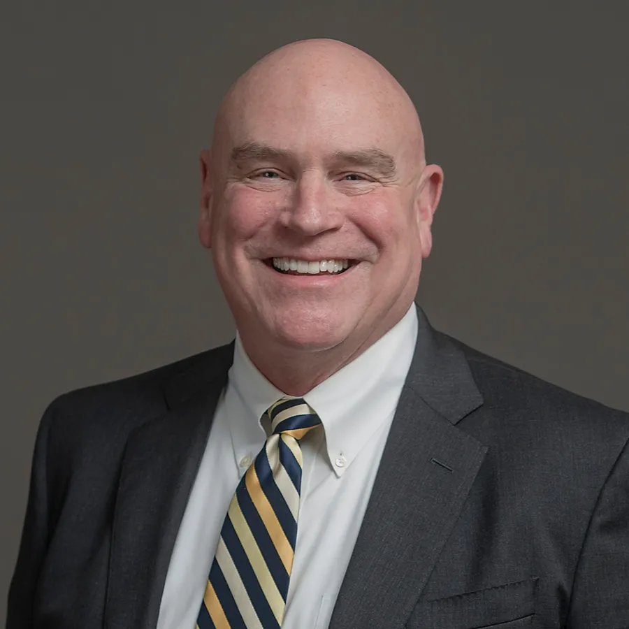 Smiling bald man in a dark suit, white shirt, and blue and yellow striped tie against gray background